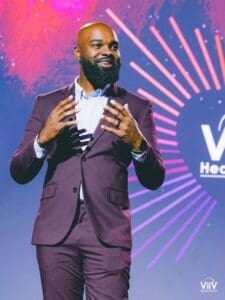 Randevyn Piérre, ViiV Healthcare Head of US External Affairs in a purple suit and white suit in front speaking in. front of the audience at USCHA